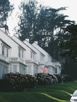 A row of white, two-story houses with gabled roofs is adorned by well-maintained gardens filled with hydrangea bushes. An American flag is prominently displayed in front of one house. Mature trees with dense foliage are visible in the background, creating a serene suburban setting. The lawn is neatly cut, and a car is partially visible on the right side of the image.