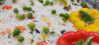 A close-up view of a colorful rice dish featuring white basmati rice mixed with bright green peas, red and yellow bell pepper slices. The rice is scattered with finely chopped herbs and vegetables, creating a vibrant and appetizing appearance.