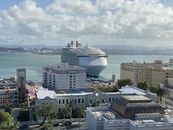 A large cruise ship is docked in a busy port area, surrounded by a range of urban buildings. The water is calm and the sky is partially cloudy, with mountains visible in the distant background. Colorful buildings and a tower are in the foreground, with greenery scattered throughout the area.