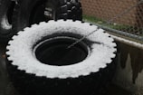 Large, heavy tires are covered with a light dusting of snow, positioned against a metal fence. A chain is attached to one of the tires. The area appears to be an industrial or outdoor setting.