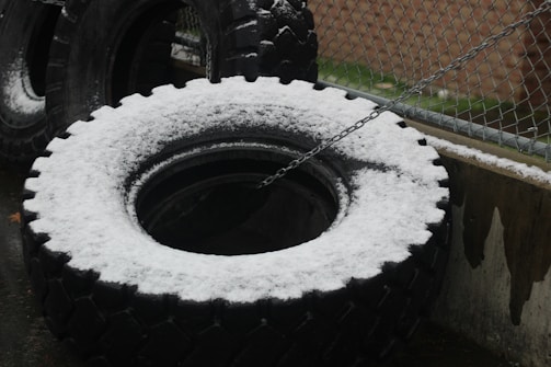 Large, heavy tires are covered with a light dusting of snow, positioned against a metal fence. A chain is attached to one of the tires. The area appears to be an industrial or outdoor setting.