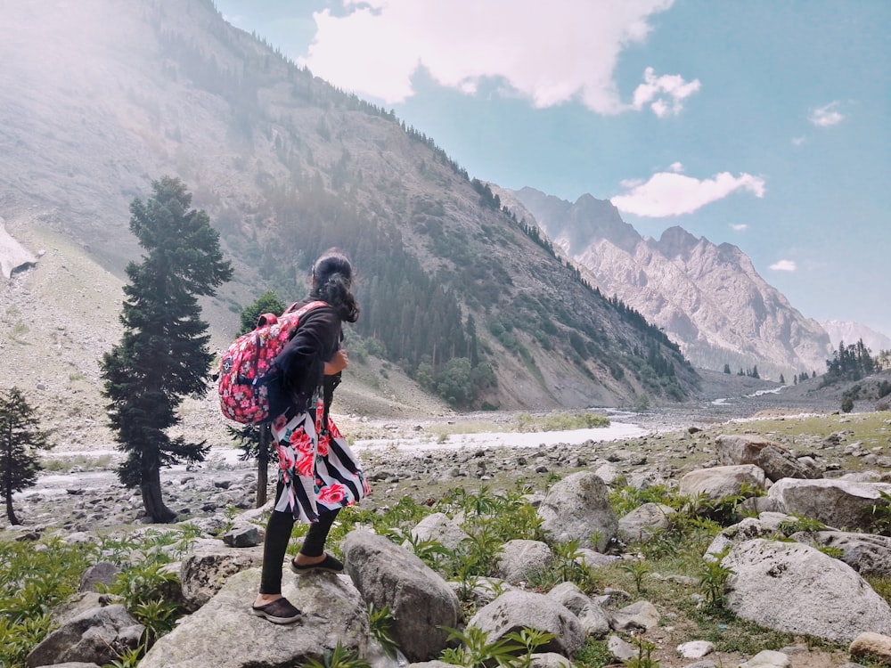 woman in red and black backpack standing on rocky ground near green trees and mountains during