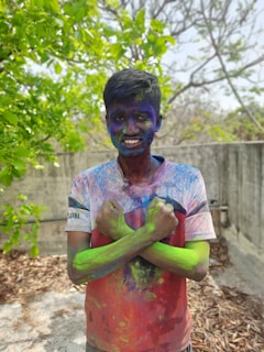 A person stands outside covered in vibrant colored powders, with a smile on their face. The background includes green trees and a concrete wall, suggesting an outdoor setting.