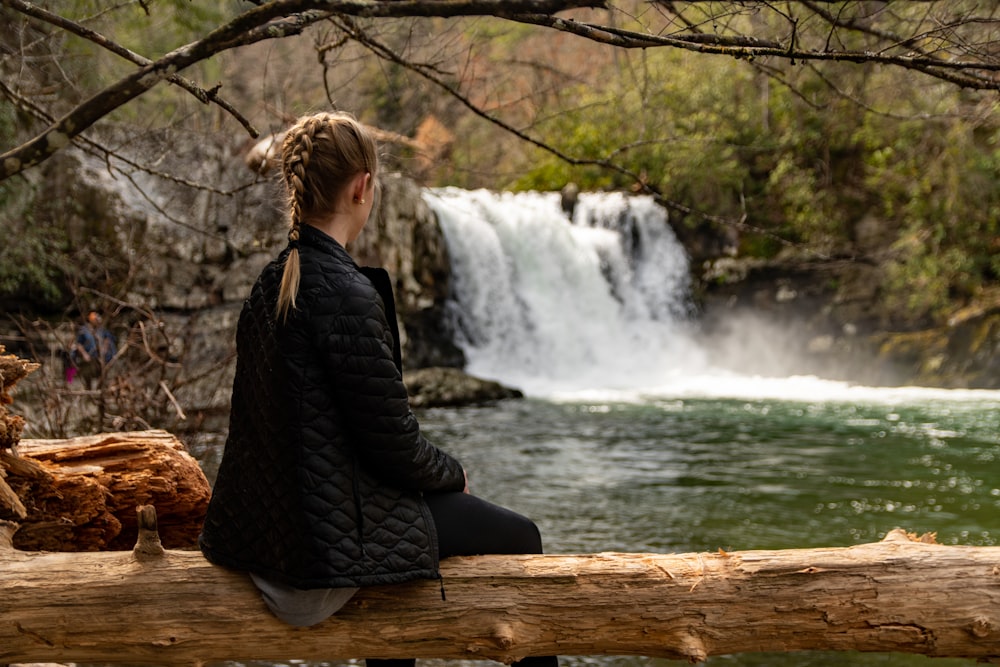 woman in black jacket sitting on brown wooden log near waterfalls during daytime