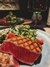 A plate with seared tuna steak, showcasing a medium rare interior with grill marks on the surface. It is accompanied by a green salad with cherry tomatoes and sprinkled sesame seeds. A glass of red wine is partially visible in the background, suggesting a dining setting.