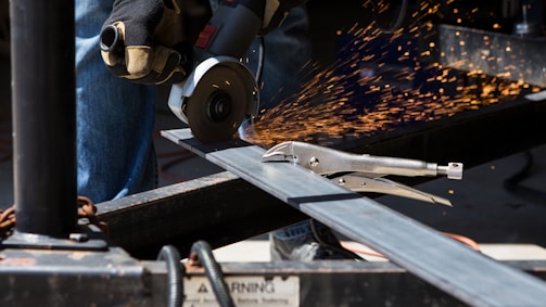 A person is using an angle grinder to cut through a metal beam, creating bright, flying sparks. They are wearing protective gloves, and a metal clamp is visible near the beam.