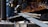A person is using an angle grinder to cut through a metal beam, creating bright, flying sparks. They are wearing protective gloves, and a metal clamp is visible near the beam.