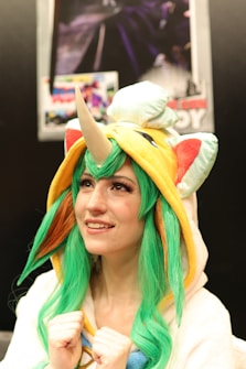 A person is wearing a colorful costume with vibrant green hair and a horned hood resembling a plush animal. The costume has orange and yellow accents, and the person is smiling while looking upwards. Behind them, a blurred poster is visible on a dark background.