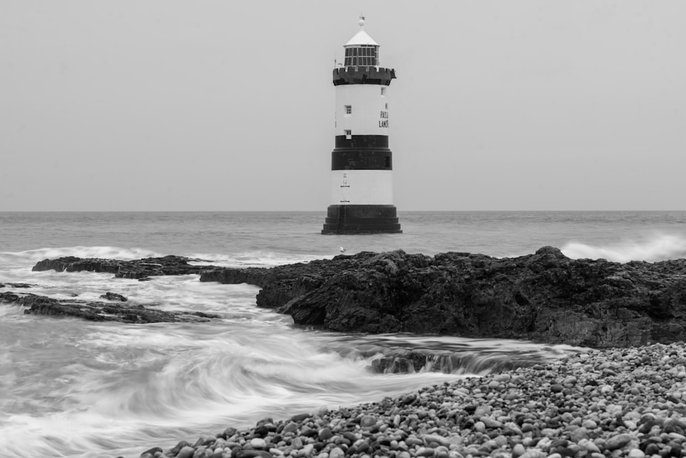 grayscale photo of lighthouse on rock formation near sea