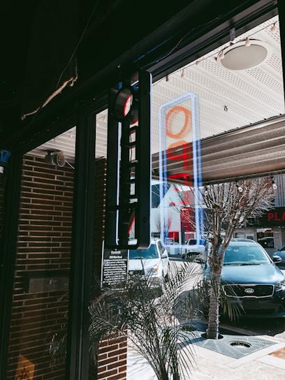 A street view from inside a building, showcasing a glass door with a blue and red open sign. Through the glass, several cars are parked along the street, including a white and a black vehicle. A neon sign is reflected on the window, and there are brick walls and decorative plants nearby.