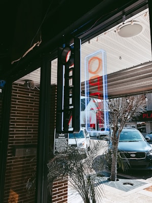 A street view from inside a building, showcasing a glass door with a blue and red open sign. Through the glass, several cars are parked along the street, including a white and a black vehicle. A neon sign is reflected on the window, and there are brick walls and decorative plants nearby.