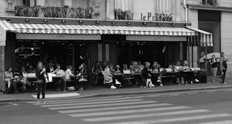 A bustling outdoor café lined with patrons sitting at tables under an awning. The café has striped awnings and flower boxes above. People are dining, chatting, and drinking coffee. Some individuals are walking along the sidewalk, and a pedestrian is crossing the street in front of the café. There are motorcycles parked and some greenery visible.