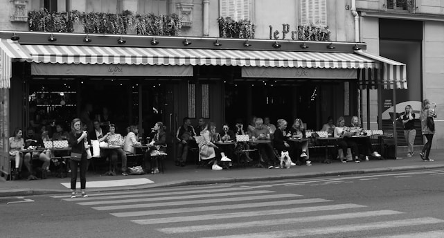 A bustling outdoor café lined with patrons sitting at tables under an awning. The café has striped awnings and flower boxes above. People are dining, chatting, and drinking coffee. Some individuals are walking along the sidewalk, and a pedestrian is crossing the street in front of the café. There are motorcycles parked and some greenery visible.