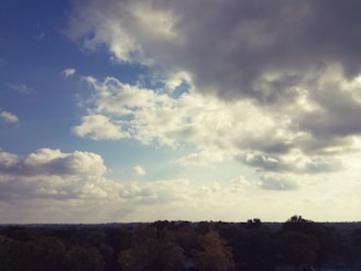 Cloud-filled sky with patches of blue peeking through, above a landscape of trees. The clouds are dense and appear to be casting shadows over the scenery below.