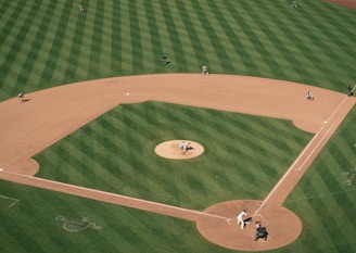 Aerial view of a baseball field showing a game in action. The pitcher is mid-throw, the batter is poised at the plate, and several players are positioned around the field. The grass is neatly mowed in a striped pattern, and the infield dirt is well-defined. The scene is dynamic, capturing a moment of play under clear skies.