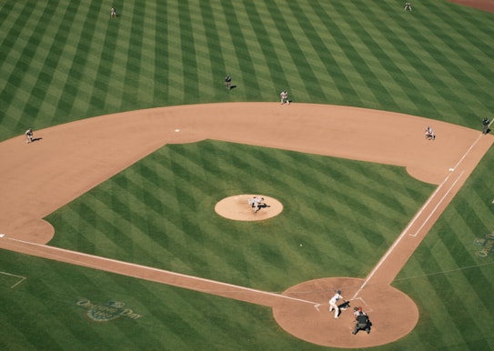 Aerial view of a baseball field showing a game in action. The pitcher is mid-throw, the batter is poised at the plate, and several players are positioned around the field. The grass is neatly mowed in a striped pattern, and the infield dirt is well-defined. The scene is dynamic, capturing a moment of play under clear skies.