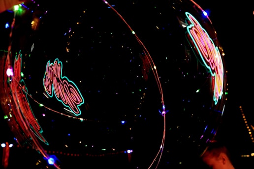 A vibrant neon sign is reflected off a transparent surface, possibly a bubble or a reflective glass. The neon colors create an abstract and dynamic visual with streaks of red, pink, and blue lights against a dark background.
