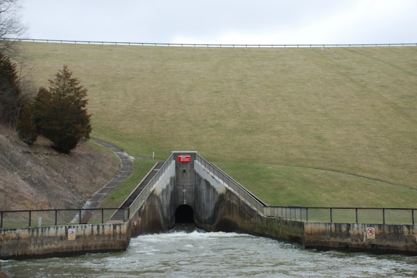 The image depicts a sloped grassy embankment with a paved pathway leading upward on the left side. At the base, there is a concrete structure resembling a spillway or culvert, with water flowing out of it into a body of water in the foreground. Metal fencing lines the top of the concrete structure, and signs can be seen attached to the fence.
