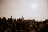 A group of people gathered in a cemetery with numerous gravestones and floral arrangements. The scene is silhouetted against a bright sky, with the spire of a church visible in the background. Tall trees and utility poles are also present.