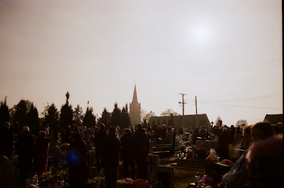 A group of people gathered in a cemetery with numerous gravestones and floral arrangements. The scene is silhouetted against a bright sky, with the spire of a church visible in the background. Tall trees and utility poles are also present.