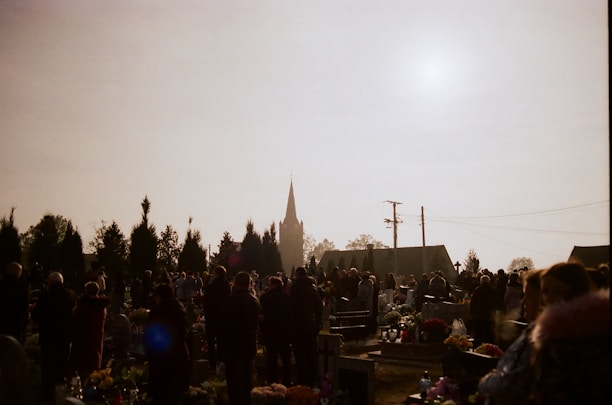 A group of people gathered in a cemetery with numerous gravestones and floral arrangements. The scene is silhouetted against a bright sky, with the spire of a church visible in the background. Tall trees and utility poles are also present.