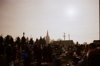 A group of people gathered in a cemetery with numerous gravestones and floral arrangements. The scene is silhouetted against a bright sky, with the spire of a church visible in the background. Tall trees and utility poles are also present.