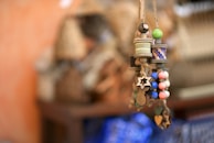 A collection of intricately designed hanging ornaments crafted from wood and thread. The ornaments include colorful beads in shades of blue, green, pink, and natural wood, along with star-shaped and heart-shaped designs. The background features a soft focus on wicker baskets and a warm orange wall.
