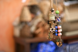 A collection of intricately designed hanging ornaments crafted from wood and thread. The ornaments include colorful beads in shades of blue, green, pink, and natural wood, along with star-shaped and heart-shaped designs. The background features a soft focus on wicker baskets and a warm orange wall.