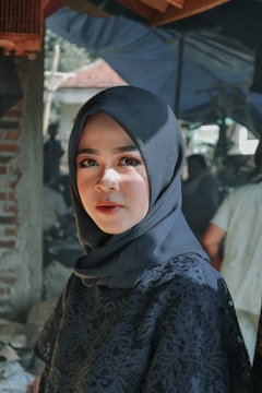 A woman wearing a dark hijab and lace outfit, standing in a sunlit environment with shadows around. She appears to be outdoors with a background of blurred greenery and an unfinished brick wall.