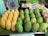 A display of ripe yellow and green mangoes is arranged on a market stall, with a traditional scale visible in the background. Nearby, packages of snacks or food items and some plastic-wrapped produce are placed.