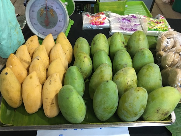 A display of ripe yellow and green mangoes is arranged on a market stall, with a traditional scale visible in the background. Nearby, packages of snacks or food items and some plastic-wrapped produce are placed.