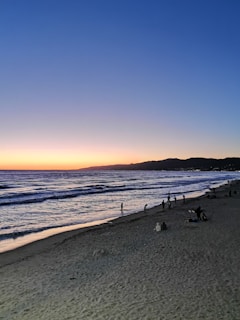 The image captures a serene beach scene at sunset, with the sky transitioning from vibrant orange near the horizon to a deep blue above. Gentle waves roll onto the sandy shore, where silhouettes of people stroll and gather in small groups. Mountains are visible in the background, adding depth to the landscape.