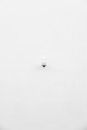 A minimalist image featuring a single security camera mounted on a stark white wall. The camera is positioned slightly off-center towards the bottom of the frame, with its small, round shape creating a subtle focal point against the expansive white background.