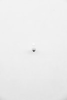 A minimalist image featuring a single security camera mounted on a stark white wall. The camera is positioned slightly off-center towards the bottom of the frame, with its small, round shape creating a subtle focal point against the expansive white background.