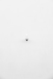 A minimalist image featuring a single security camera mounted on a stark white wall. The camera is positioned slightly off-center towards the bottom of the frame, with its small, round shape creating a subtle focal point against the expansive white background.