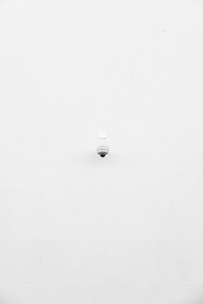 A minimalist image featuring a single security camera mounted on a stark white wall. The camera is positioned slightly off-center towards the bottom of the frame, with its small, round shape creating a subtle focal point against the expansive white background.