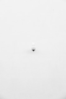 A minimalist image featuring a single security camera mounted on a stark white wall. The camera is positioned slightly off-center towards the bottom of the frame, with its small, round shape creating a subtle focal point against the expansive white background.