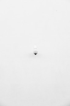 A minimalist image featuring a single security camera mounted on a stark white wall. The camera is positioned slightly off-center towards the bottom of the frame, with its small, round shape creating a subtle focal point against the expansive white background.