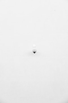 A minimalist image featuring a single security camera mounted on a stark white wall. The camera is positioned slightly off-center towards the bottom of the frame, with its small, round shape creating a subtle focal point against the expansive white background.