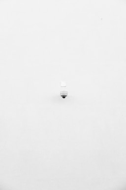 A minimalist image featuring a single security camera mounted on a stark white wall. The camera is positioned slightly off-center towards the bottom of the frame, with its small, round shape creating a subtle focal point against the expansive white background.