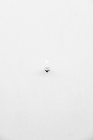 A minimalist image featuring a single security camera mounted on a stark white wall. The camera is positioned slightly off-center towards the bottom of the frame, with its small, round shape creating a subtle focal point against the expansive white background.