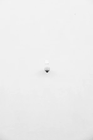 A minimalist image featuring a single security camera mounted on a stark white wall. The camera is positioned slightly off-center towards the bottom of the frame, with its small, round shape creating a subtle focal point against the expansive white background.