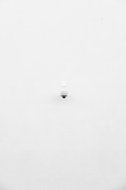 A minimalist image featuring a single security camera mounted on a stark white wall. The camera is positioned slightly off-center towards the bottom of the frame, with its small, round shape creating a subtle focal point against the expansive white background.