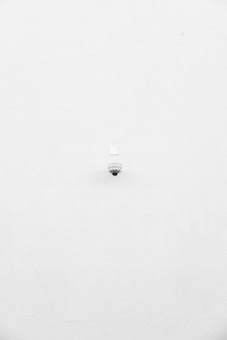 A minimalist image featuring a single security camera mounted on a stark white wall. The camera is positioned slightly off-center towards the bottom of the frame, with its small, round shape creating a subtle focal point against the expansive white background.