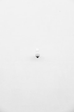 A minimalist image featuring a single security camera mounted on a stark white wall. The camera is positioned slightly off-center towards the bottom of the frame, with its small, round shape creating a subtle focal point against the expansive white background.