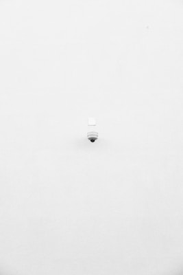 A minimalist image featuring a single security camera mounted on a stark white wall. The camera is positioned slightly off-center towards the bottom of the frame, with its small, round shape creating a subtle focal point against the expansive white background.