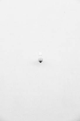 A minimalist image featuring a single security camera mounted on a stark white wall. The camera is positioned slightly off-center towards the bottom of the frame, with its small, round shape creating a subtle focal point against the expansive white background.