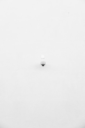 A minimalist image featuring a single security camera mounted on a stark white wall. The camera is positioned slightly off-center towards the bottom of the frame, with its small, round shape creating a subtle focal point against the expansive white background.