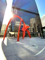 A striking red metal sculpture stands prominently in front of a tall, modern glass building. The sun shines brightly, creating lens flares and casting long shadows on the pavement. The surrounding area is an open plaza with additional skyscrapers visible in the background.