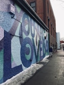 A colorful mural with large letters spelling out an inspirational message adorns the side of a brick building. The mural uses shades of blue, green, and purple. Snow lines the edge of the sidewalk, and a person wearing a blue jacket and hat is walking along the path.